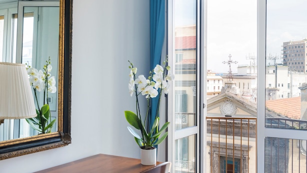 A room with a large window, a mirror, a wooden table, and a potted orchid. - Fiorentini Residence (Naples)