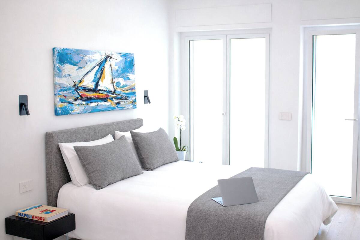A modern bedroom with a bed, a laptop, and a painting of a sailboat.