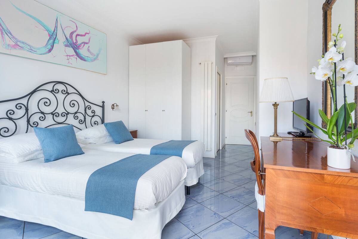 A hotel room with two beds, a wooden desk, a mirror, and a painting on the wall.