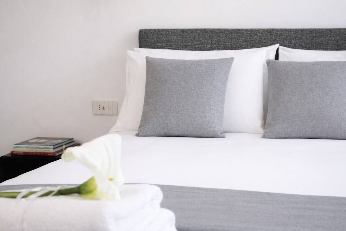 A neatly made bed with grey and white pillows, a white bedside table with books, and a white flower on a white pillow.
