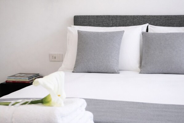 Una cama bien hecha con almohadas grises y blancas, una mesita de noche blanca con libros y una flor blanca sobre una almohada blanca.