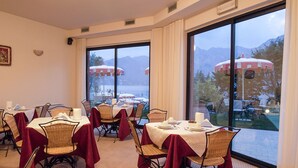 A dining area with tables set for four, large windows with a view of mountains and umbrellas outside. - Hotel Oasi Beach (Malcesine)