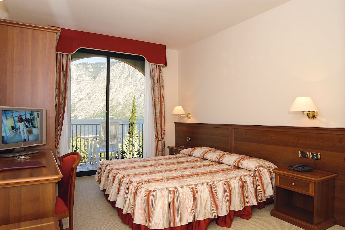 A hotel room with a large bed, a desk with a television, a nightstand, and a balcony with a view of the mountains.