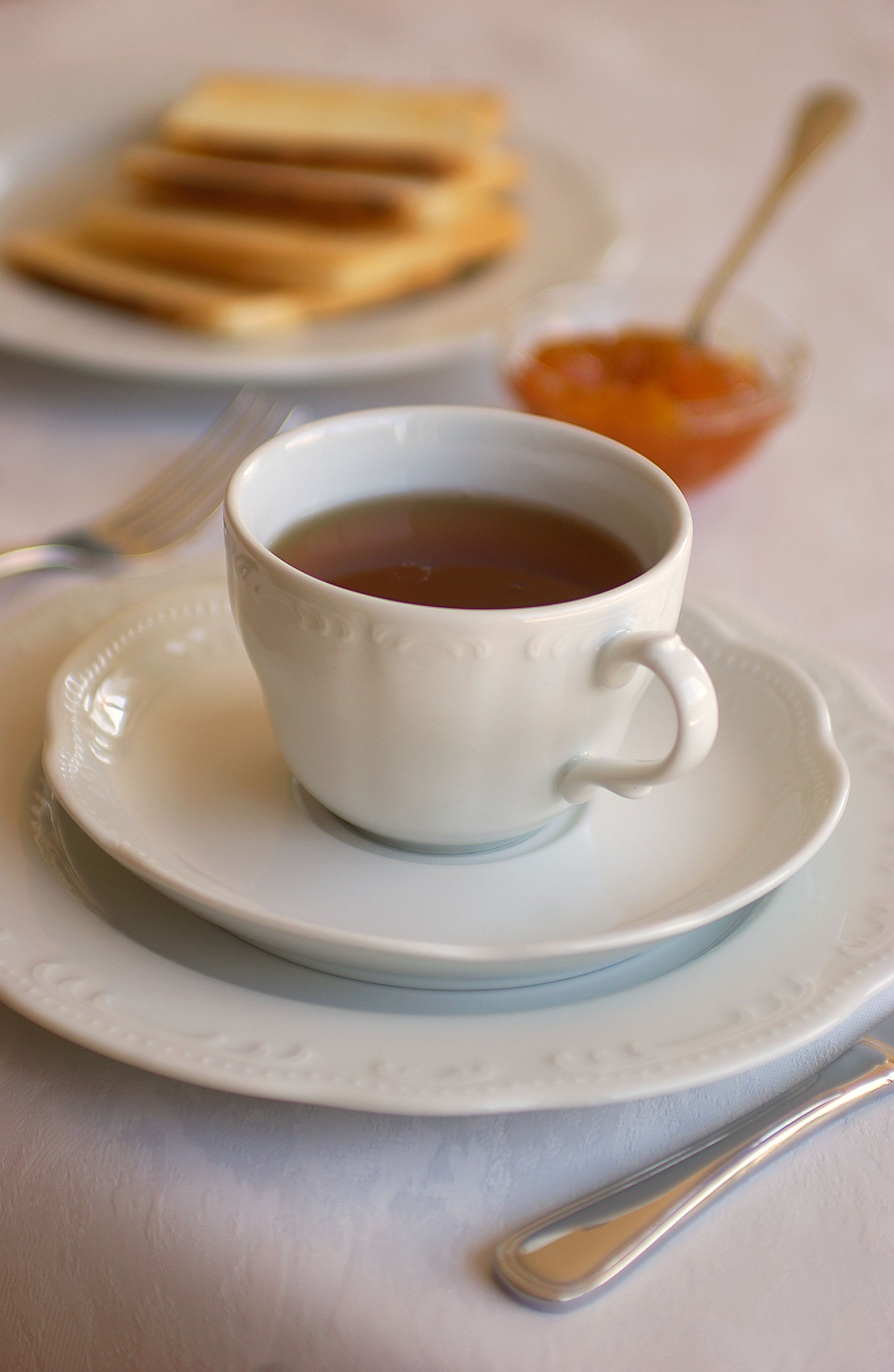 A cup of tea on a saucer with toast and jam in the background.