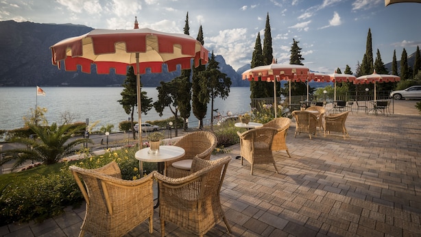 An outdoor patio with wicker furniture and red and white striped umbrellas overlooking a lake. - Hotel Oasi Beach (Malcesine)