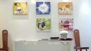 Un'area di accoglienza con una scrivania bianca, due sedie di legno e quattro opere d'arte appese al muro.