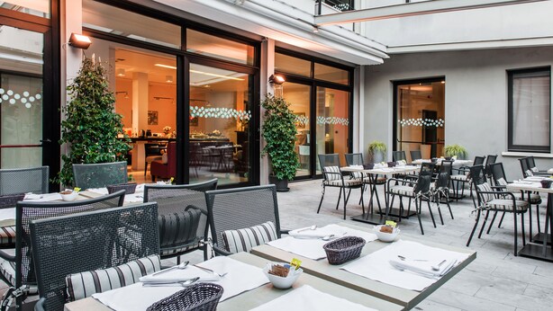 An outdoor dining area with wicker furniture, potted plants, and a view of the interior restaurant. - Hotel Casa Poli (Mantua)