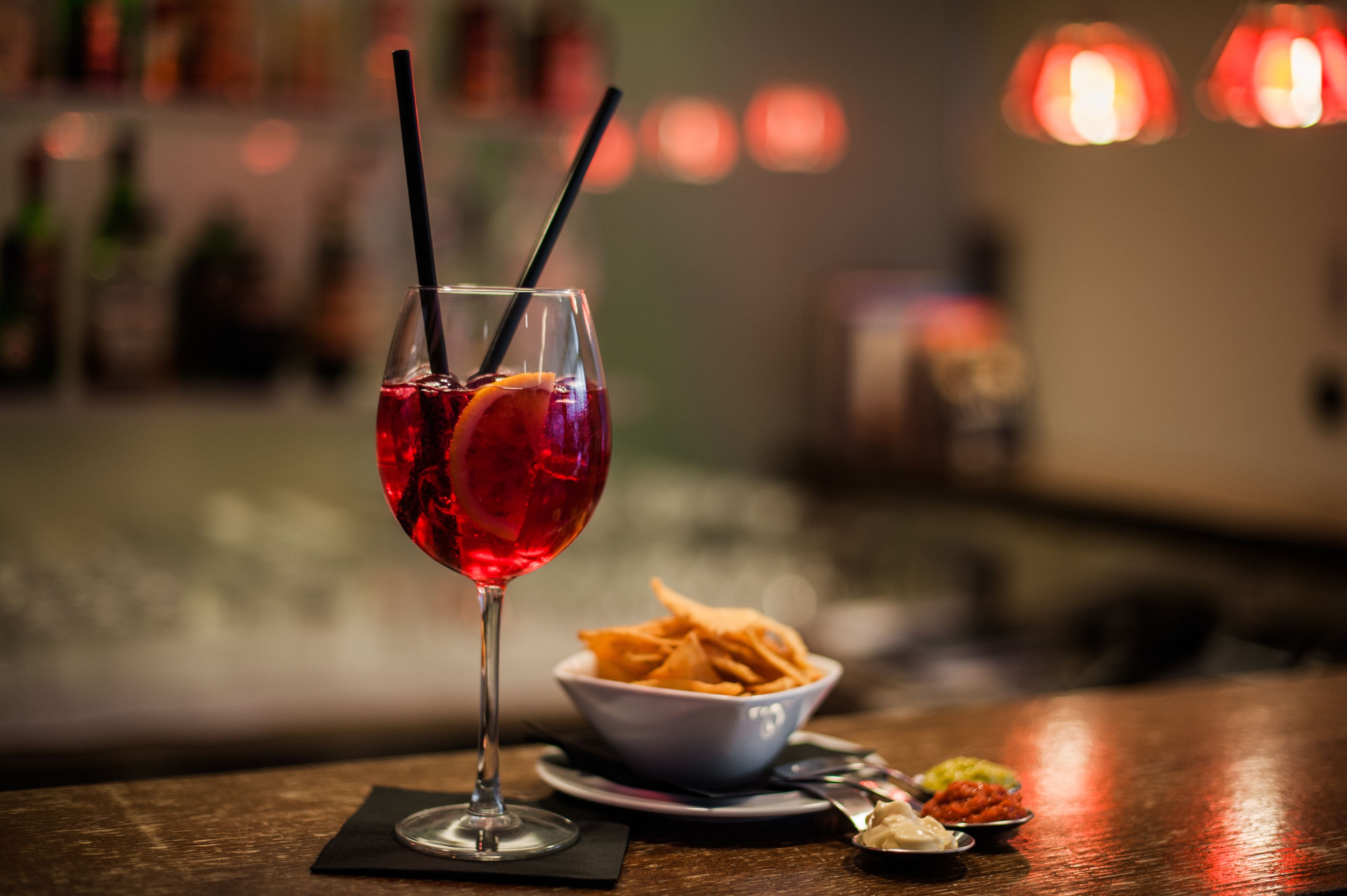 A glass of red beverage with straws, a bowl of fries, and a spoon on a table.