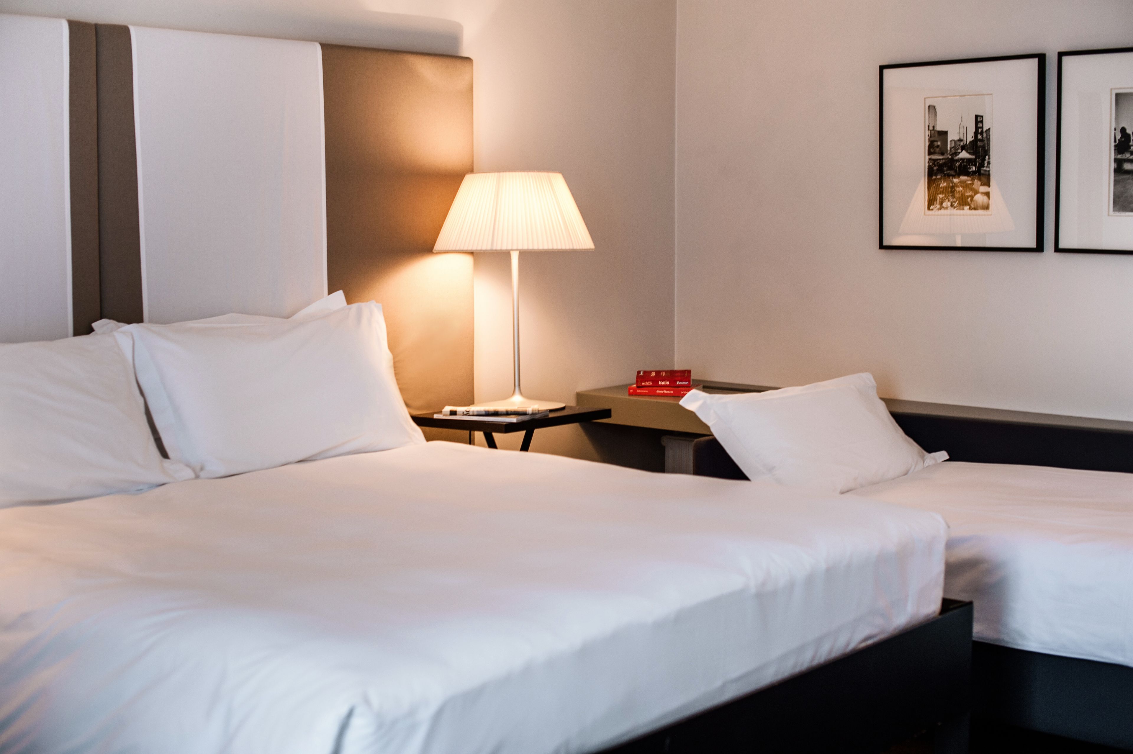 A hotel room with two beds, a bedside table with a lamp, and two framed pictures on the wall.