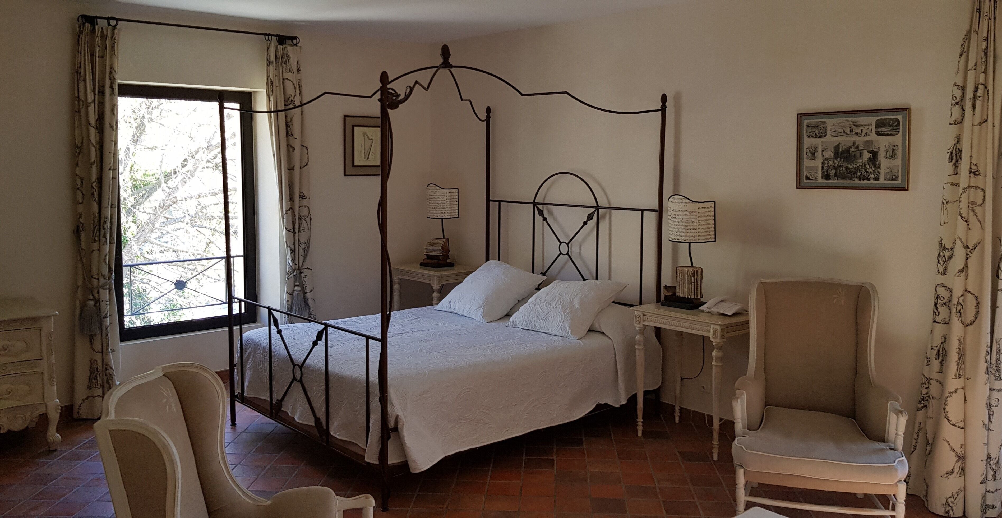 A four-poster bed with a white bedspread, a bedside table with a lamp, a chair, and a window with curtains.