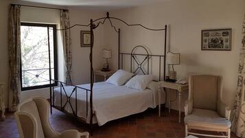 A four-poster bed with a white bedspread, a bedside table with a lamp, a chair, and a window with curtains.