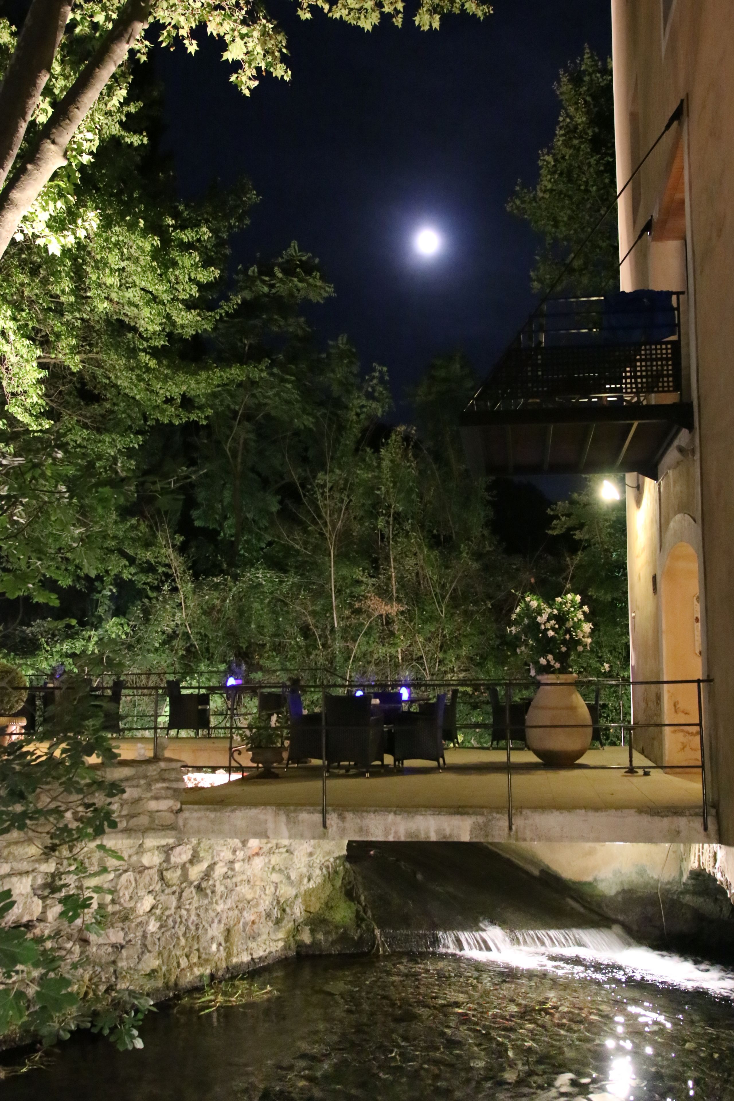 A nighttime outdoor area with a small waterfall, stone walls, and a wooden deck with seating.