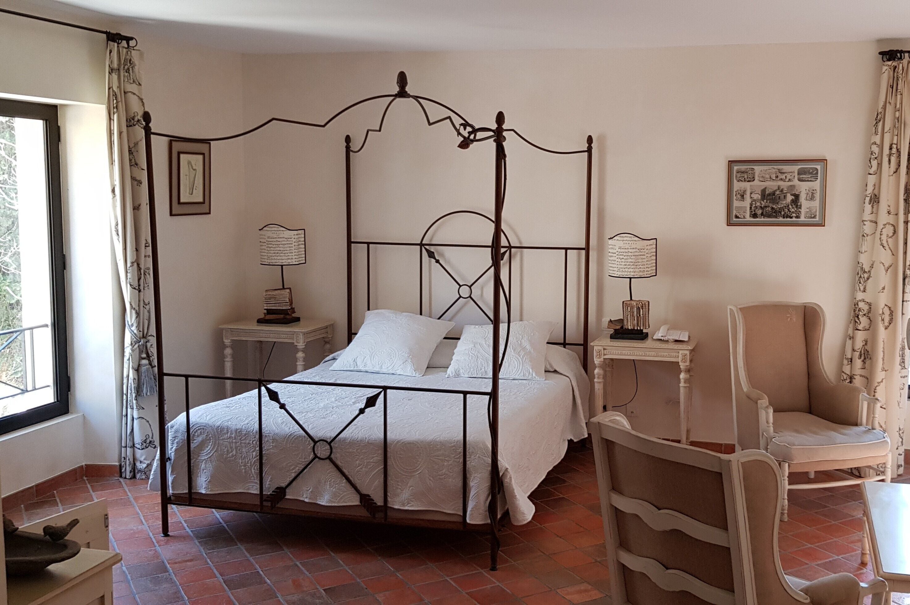 Una cama con dosel, un edredón blanco, dos mesitas de noche con lámparas, una silla y una pequeña mesa con un libro.