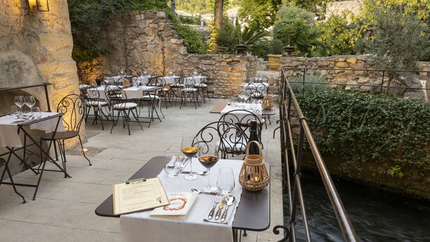 An outdoor dining area with stone walls and a view of a river. Tables are set with white tablecloths, metal chairs, and various dining utensils.