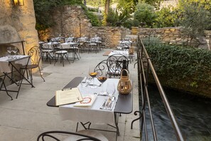 Un espace de restauration en plein air, avec des murs en pierre et une vue sur une rivière. Les tables sont dressées avec des nappes blanches, des chaises en métal et divers couverts.