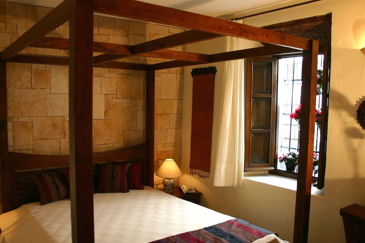 A four-poster bed with a stone wall backdrop, a bedside lamp, and a window with curtains.