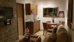 Una sala de estar con un sofá, una mesa de centro con un florero, un televisor montado en una pared de piedra, una cocina con armarios y un fregadero, y una mesa de comedor con sillas.