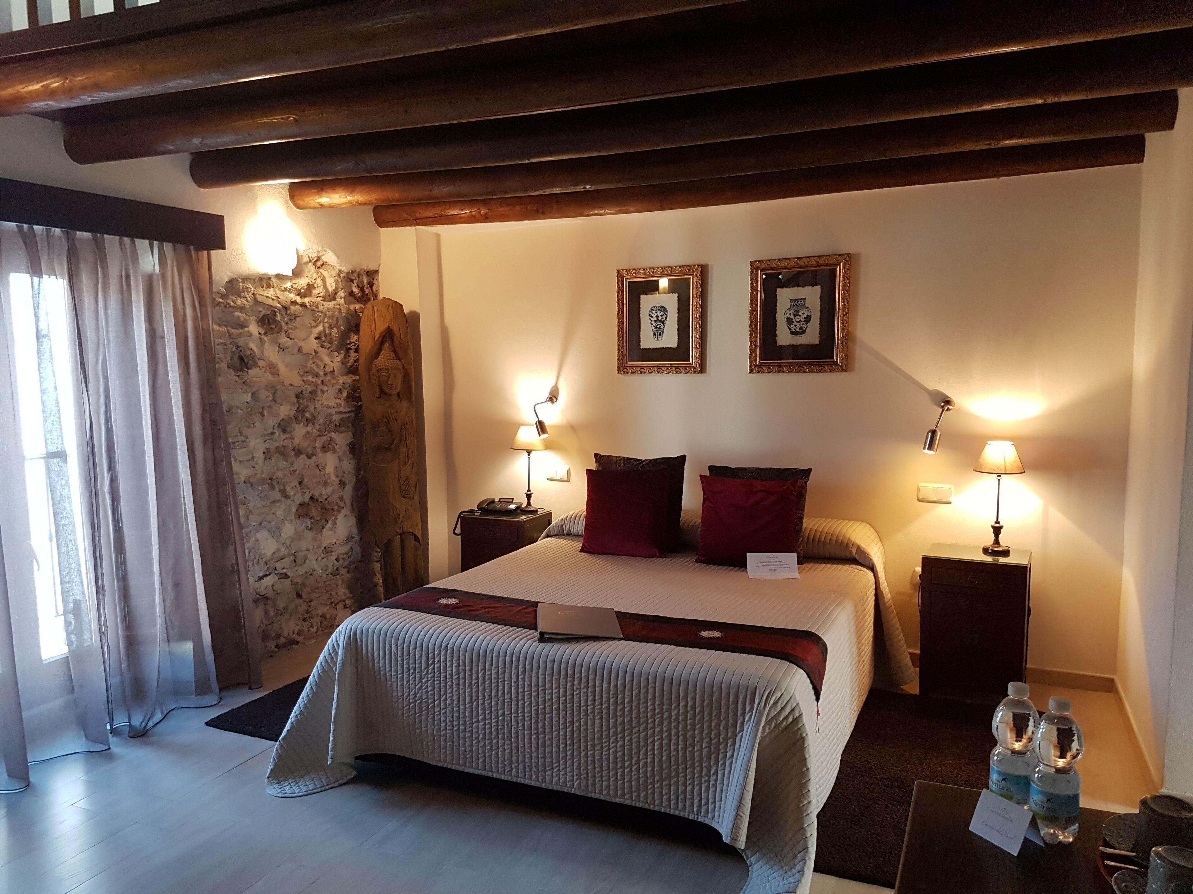 Ett hotellrum med en säng, två sänglampor, en träpanel vid sänggaveln, en vägg av sten och en stor Buddha-staty.