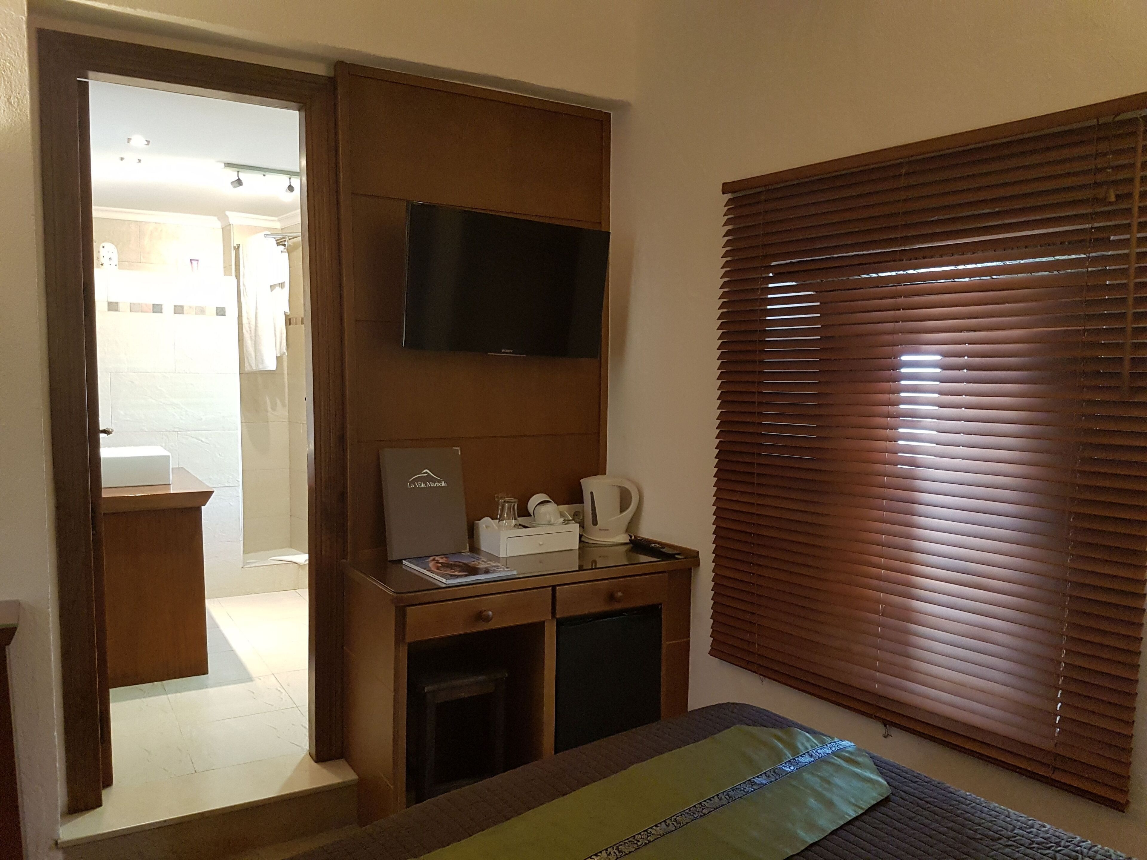 Una habitación de hotel con una cama, un televisor montado en la pared, un escritorio con cafetera y una ventana con persianas.