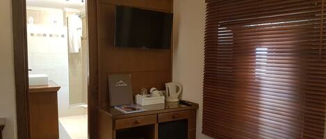 A hotel room with a bed, a TV mounted on the wall, a desk with a coffee maker, and a window with blinds.