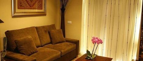 Una sala de estar con un sofá marrón, una mesa de centro de madera con una orquídea rosa y un ventanal con persianas verticales.