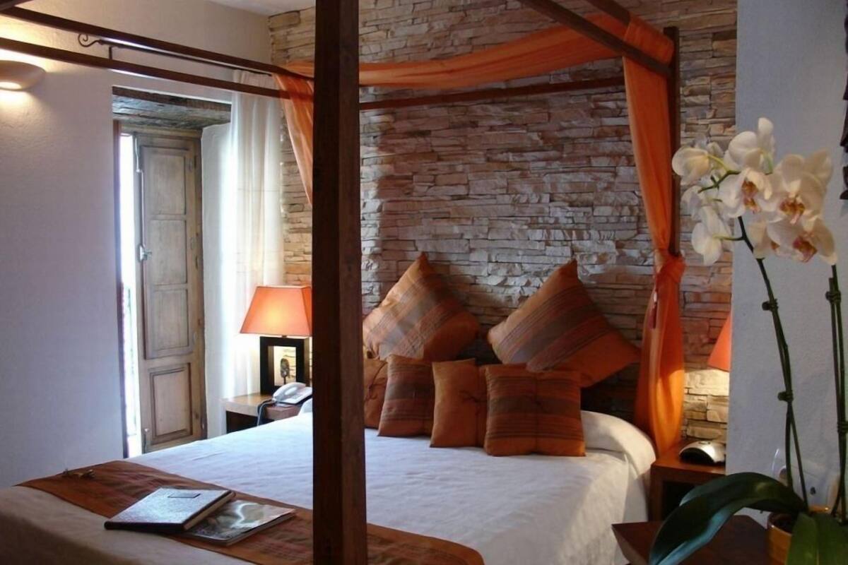 A four-poster bed with a stone wall backdrop, a bedside table with a lamp, and a vase with white orchids.
