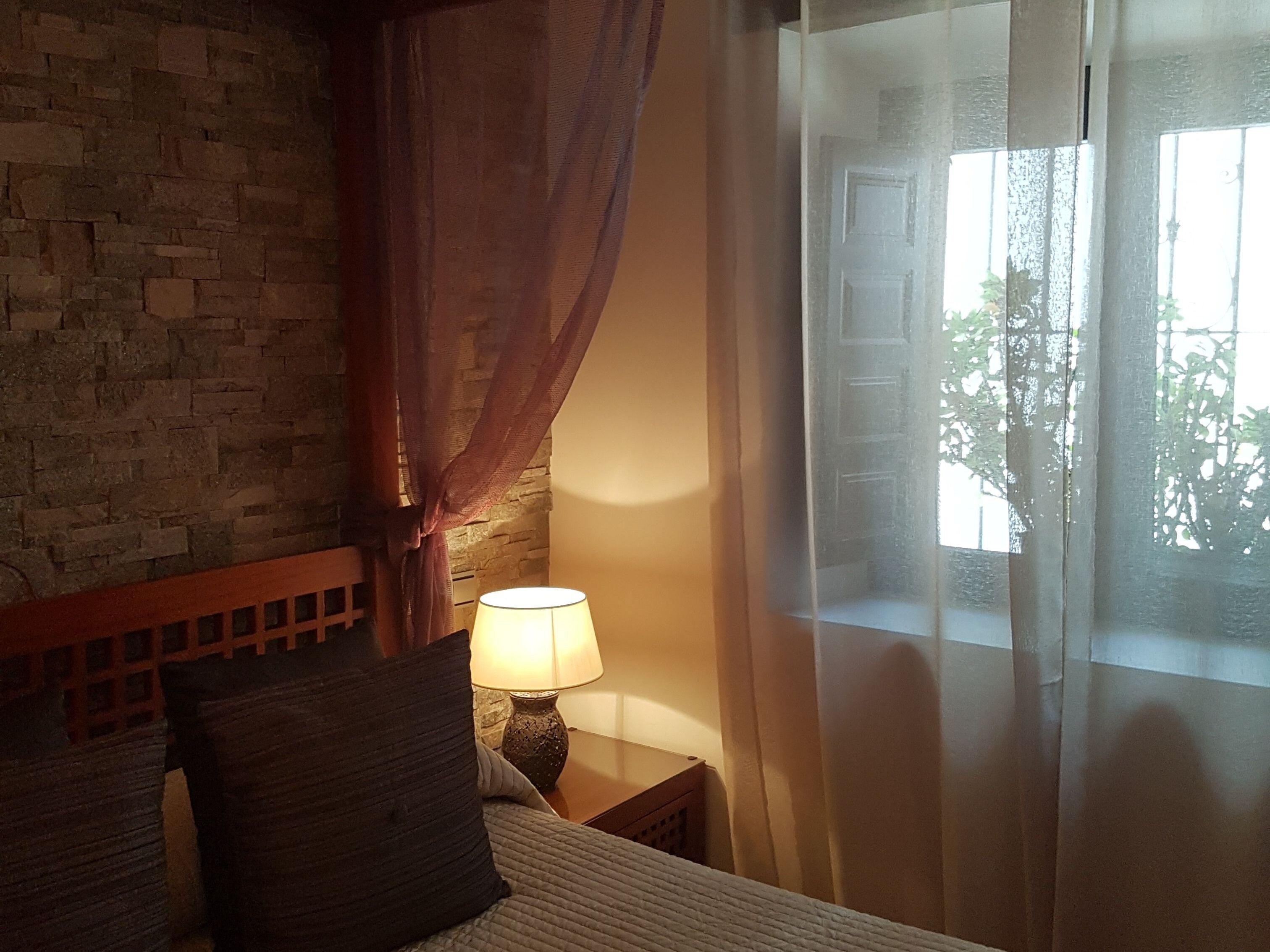 A bedroom with a stone wall, a bed with a headboard, a nightstand with a lamp, and a window with sheer curtains.