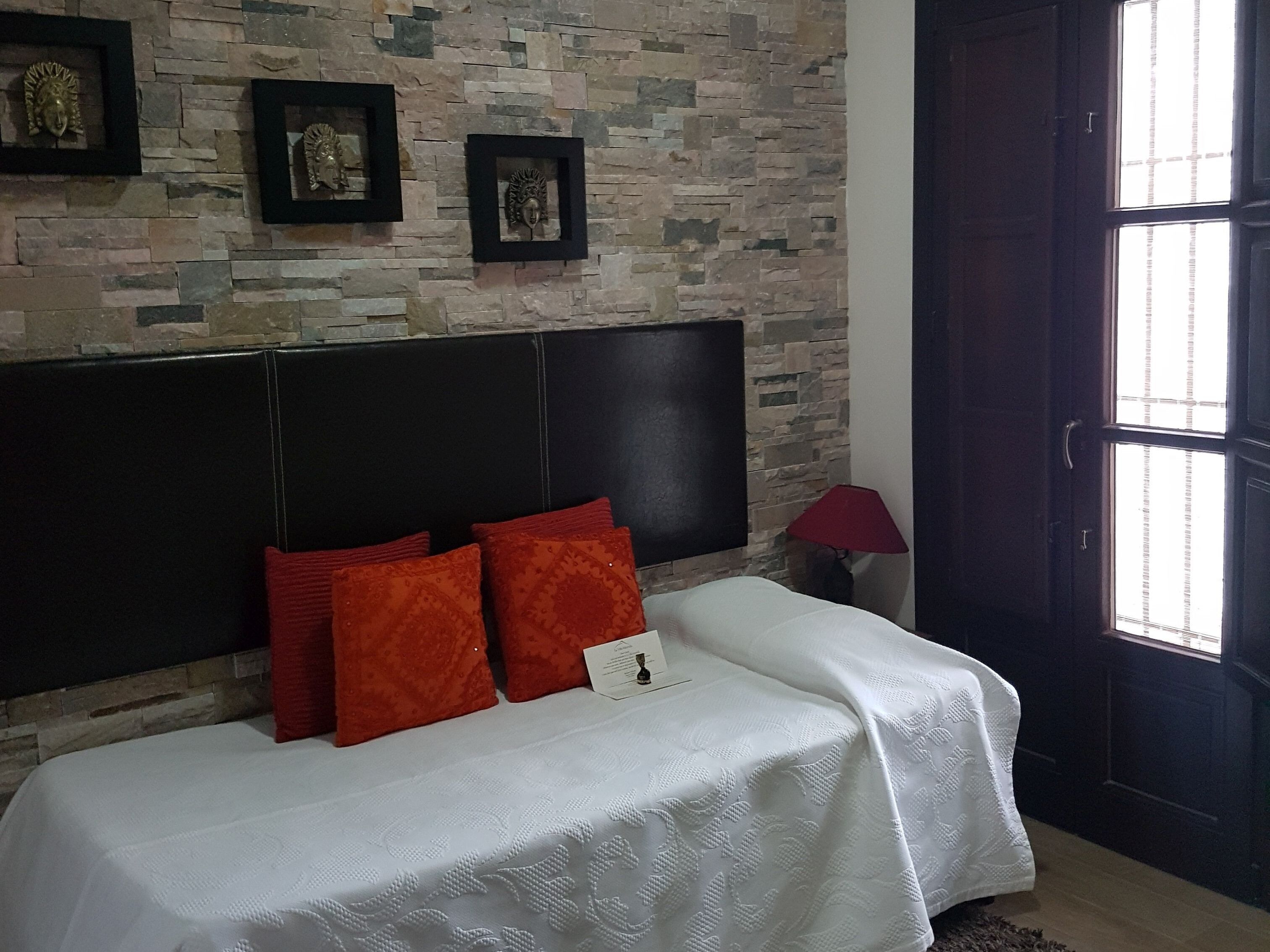 A bedroom with a stone wall, a black headboard, a white bedspread, and red pillows.