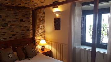A bedroom with a stone wall, a wooden headboard, a bedside lamp, a window with curtains, and a view of greenery outside.
