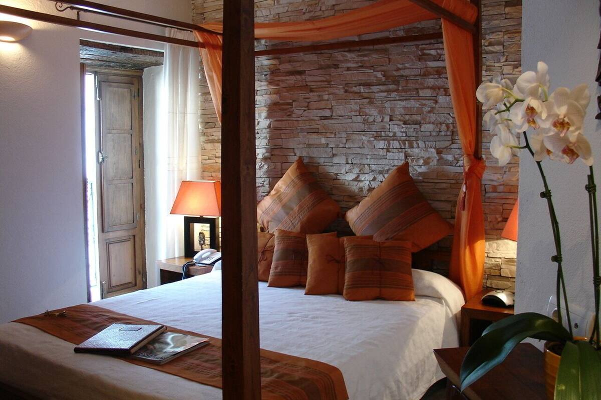 A bedroom with a four-poster bed, a stone wall, and a nightstand with a lamp.