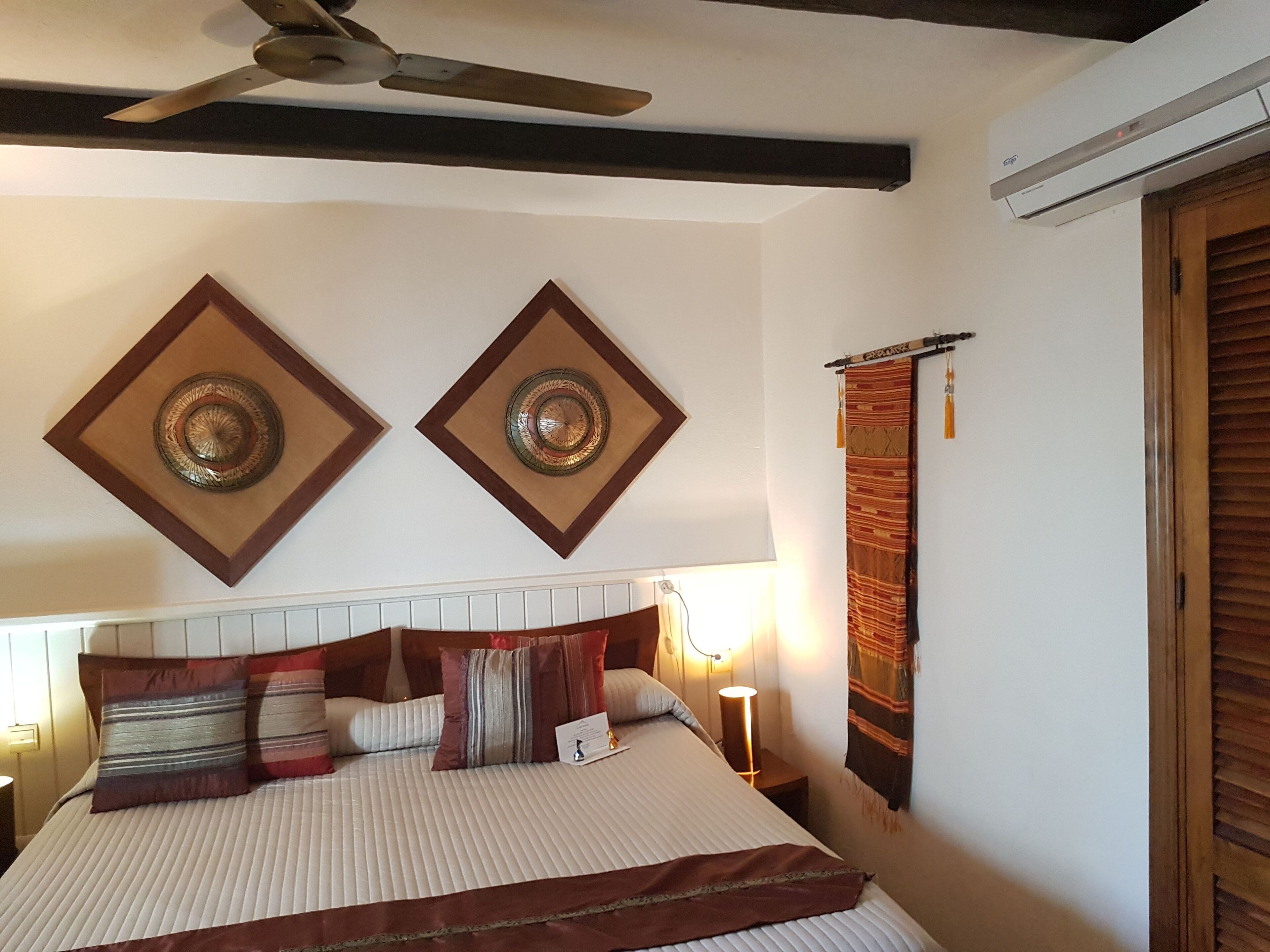 Une chambre à coucher avec un lit, un ventilateur de plafond, des œuvres d’art décoratives au mur et une fenêtre avec des rideaux.