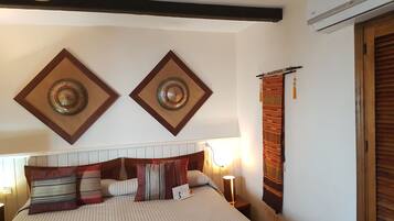 Une chambre à coucher avec un lit, un ventilateur de plafond, des œuvres d’art décoratives au mur et une fenêtre avec des rideaux.