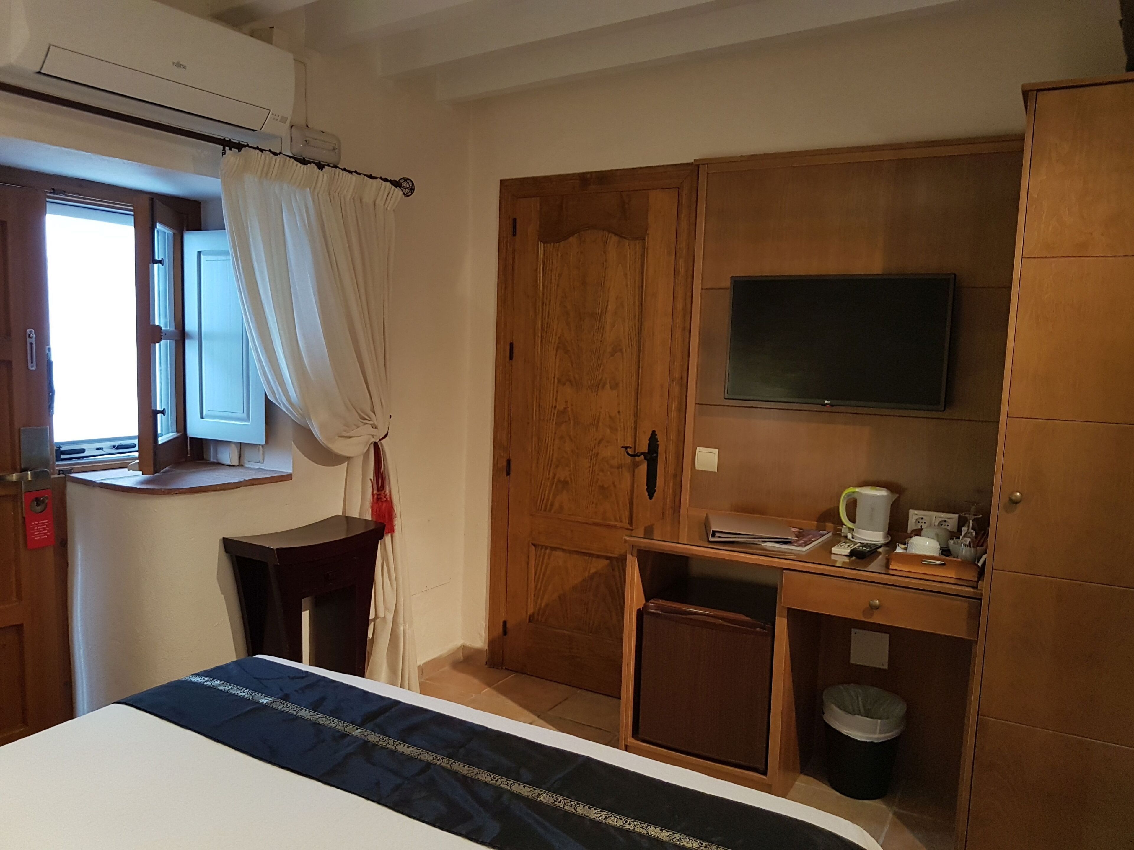 A bedroom with a bed, a desk with a TV, a wardrobe, and a window with curtains.