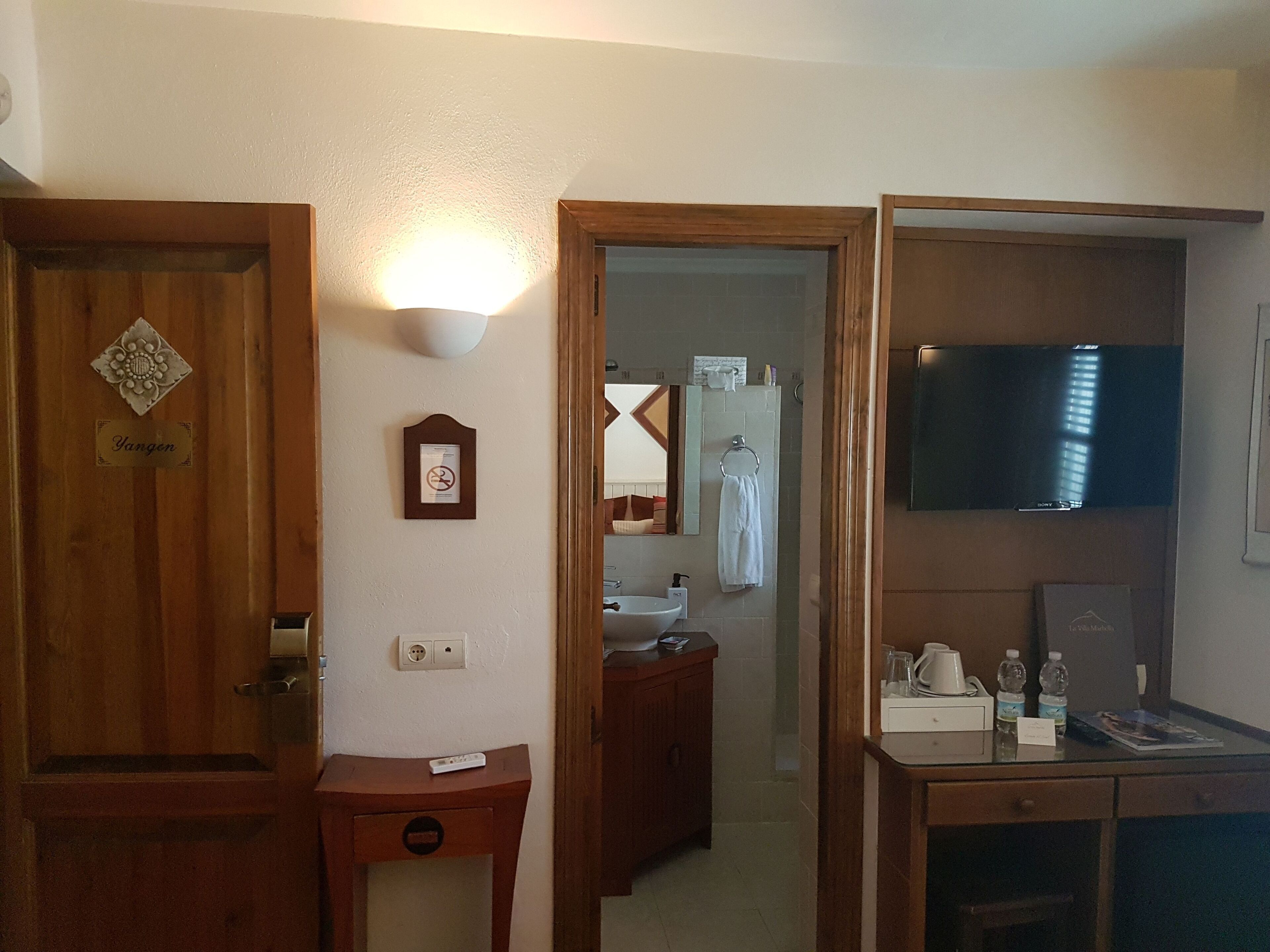 A hotel bathroom with a wooden door, a wall-mounted light, a framed sign, a mirror, a towel rack, a sink, and a television.