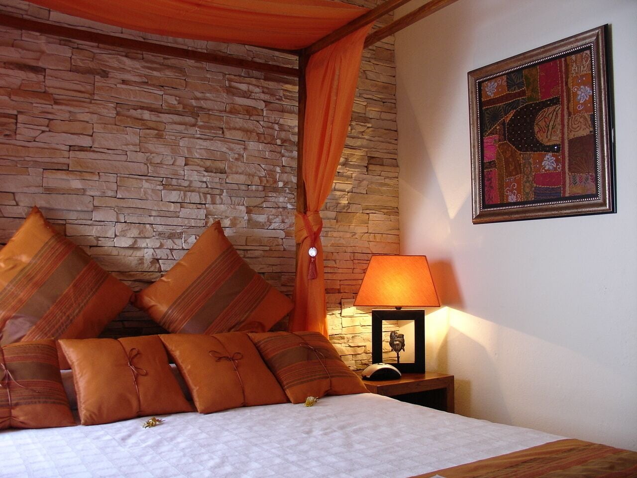 Une chambre avec un mur en pierre, un lit recouvert de plusieurs coussins, une table de chevet avec une lampe et une œuvre d’art encadrée accrochée au mur.