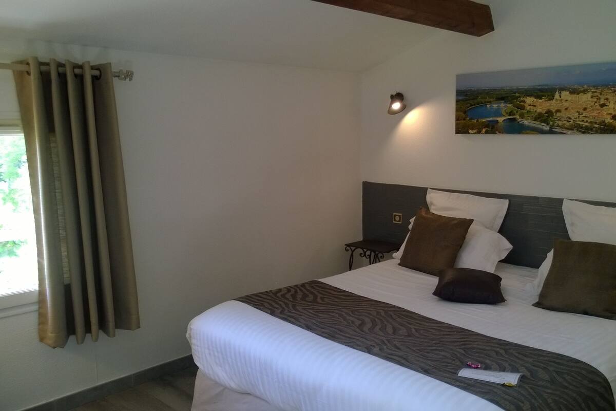 Un lit bien fait, agrémenté d’un couvre-lit à motifs, une table de chevet, une lampe fixée au mur et un tableau représentant un paysage.