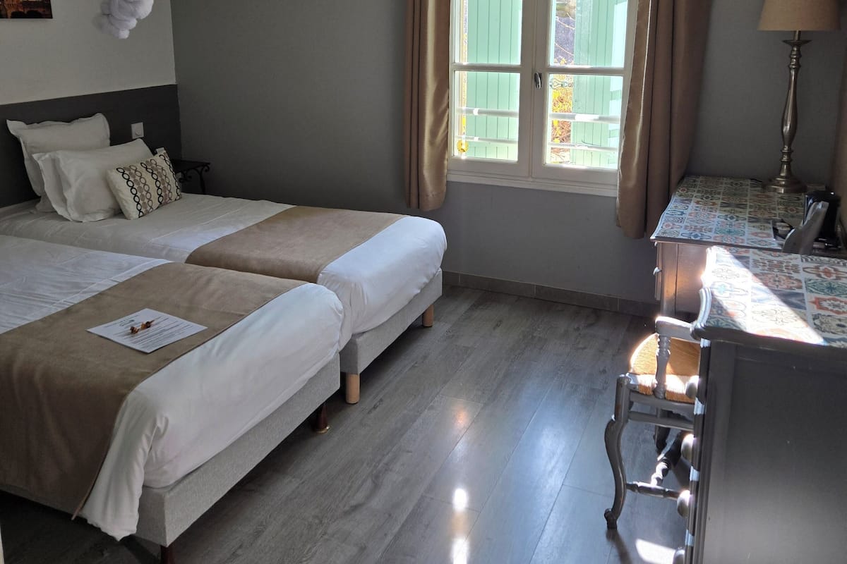 Une chambre d’hôtel avec deux lits, un bureau, une chaise, une fenêtre avec des rideaux, une moustiquaire suspendue et une lampe.