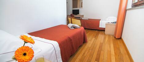 Une chambre d’hôtel avec deux lits, un plancher en bois, une petite table ornée d’un vase de fleurs, un verre de jus de fruits et un panier de fruits.