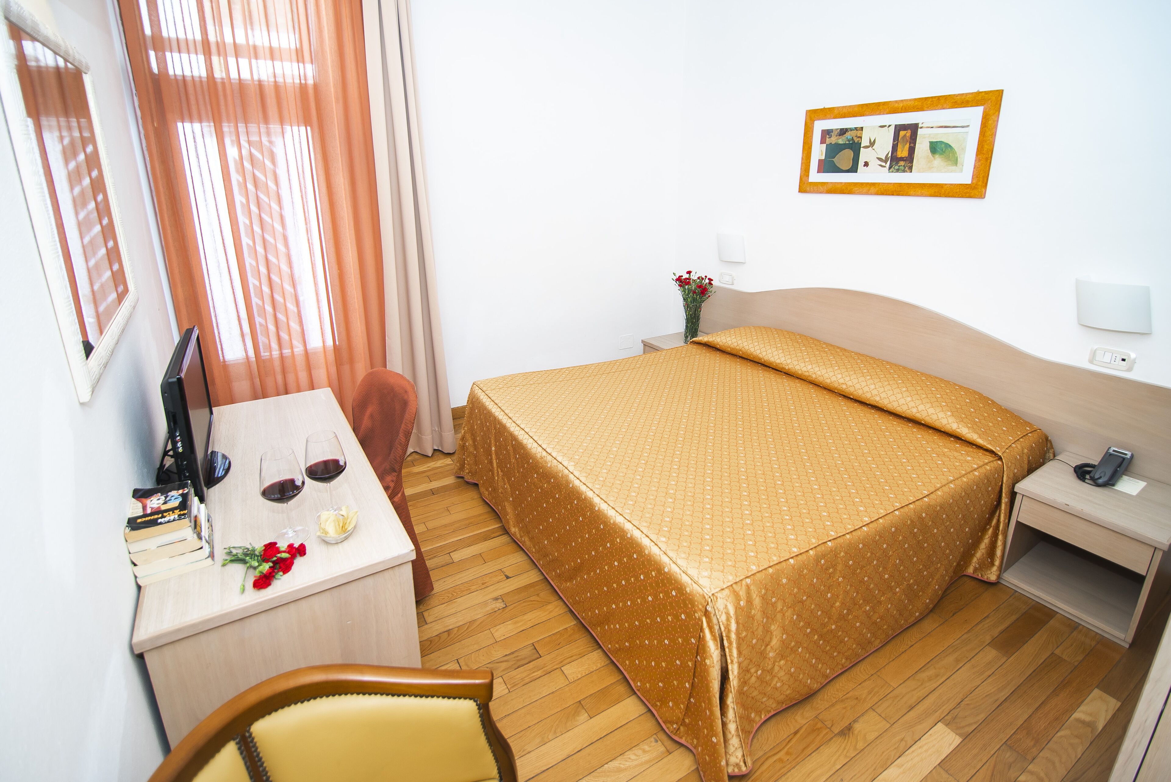 Habitación de hotel con cama, escritorio con computadora, una silla, mesita de noche y un cuadro en la pared.