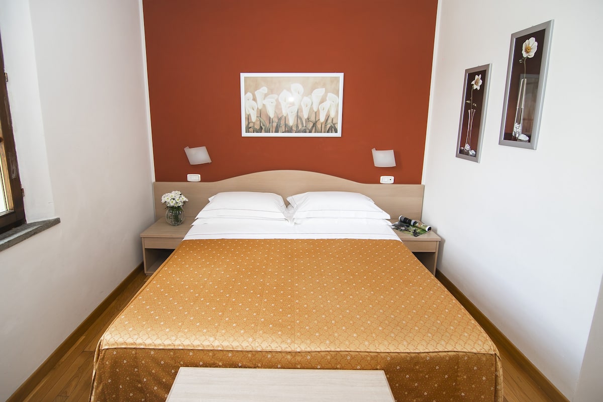 A neatly made bed with a brown polka dot bedspread, a white pillow, and a white duvet. There is a nightstand with a vase of flowers and a lamp. The room has a red accent wall and a framed artwork above the bed.