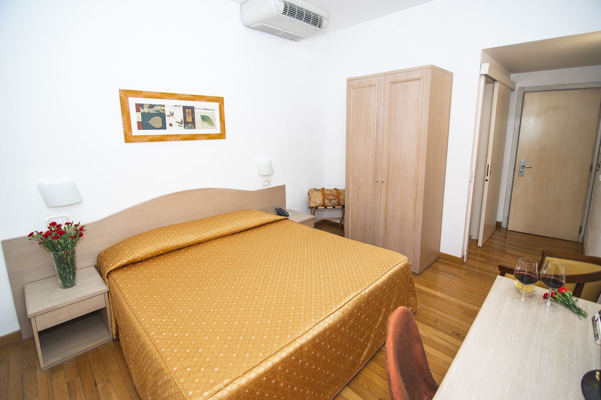 A hotel room with a bed, a desk with two glasses of wine, a vase with flowers, and a framed picture on the wall.