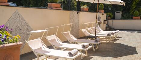 A row of sun loungers with towels, a large umbrella, and potted plants in a patio area.
