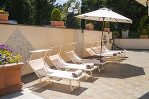A row of sun loungers with towels, a large umbrella, and potted plants in a patio area. - Hotel La Colonna (Siena)