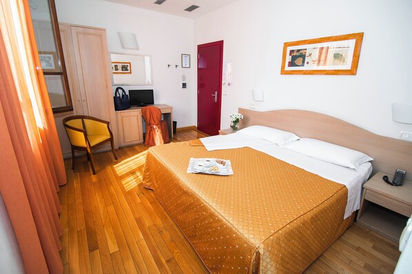 A hotel room with a bed, a desk with a computer, a chair, a red door, and a framed picture on the wall. - Hotel La Colonna (Siena)