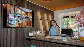 A hotel reception desk with a TV displaying a breakfast buffet, a computer, and a vase of flowers. - Hotel Burgschmiet (Nuremberg)