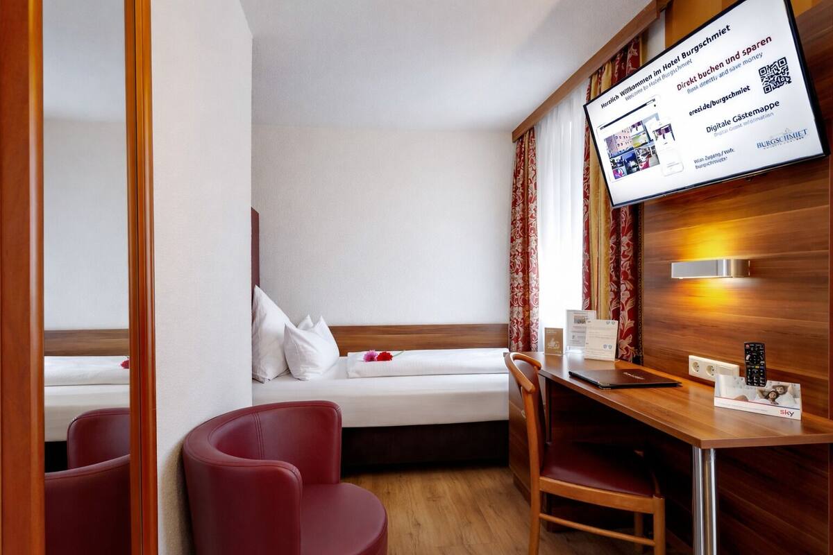 A hotel room with a bed, a desk, a chair, a TV mounted on the wall, and a window with curtains.