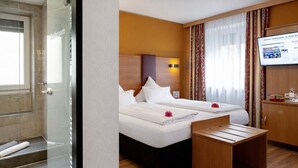A hotel room with a shower, two beds, a wooden bench, a television, and a desk with toiletries. - Hotel Burgschmiet (Nuremberg)