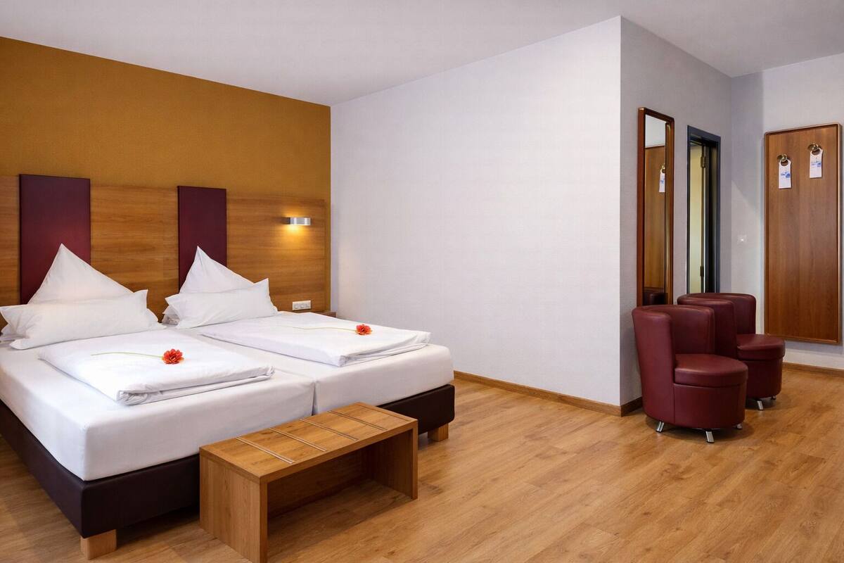 A hotel room with two beds, a wooden bench, a seating area with red chairs, and a mirror on the wall.