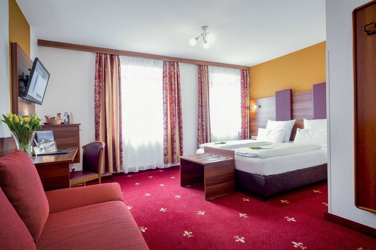 A hotel room with a bed, a desk with a computer, a chair, a small table, and a TV.