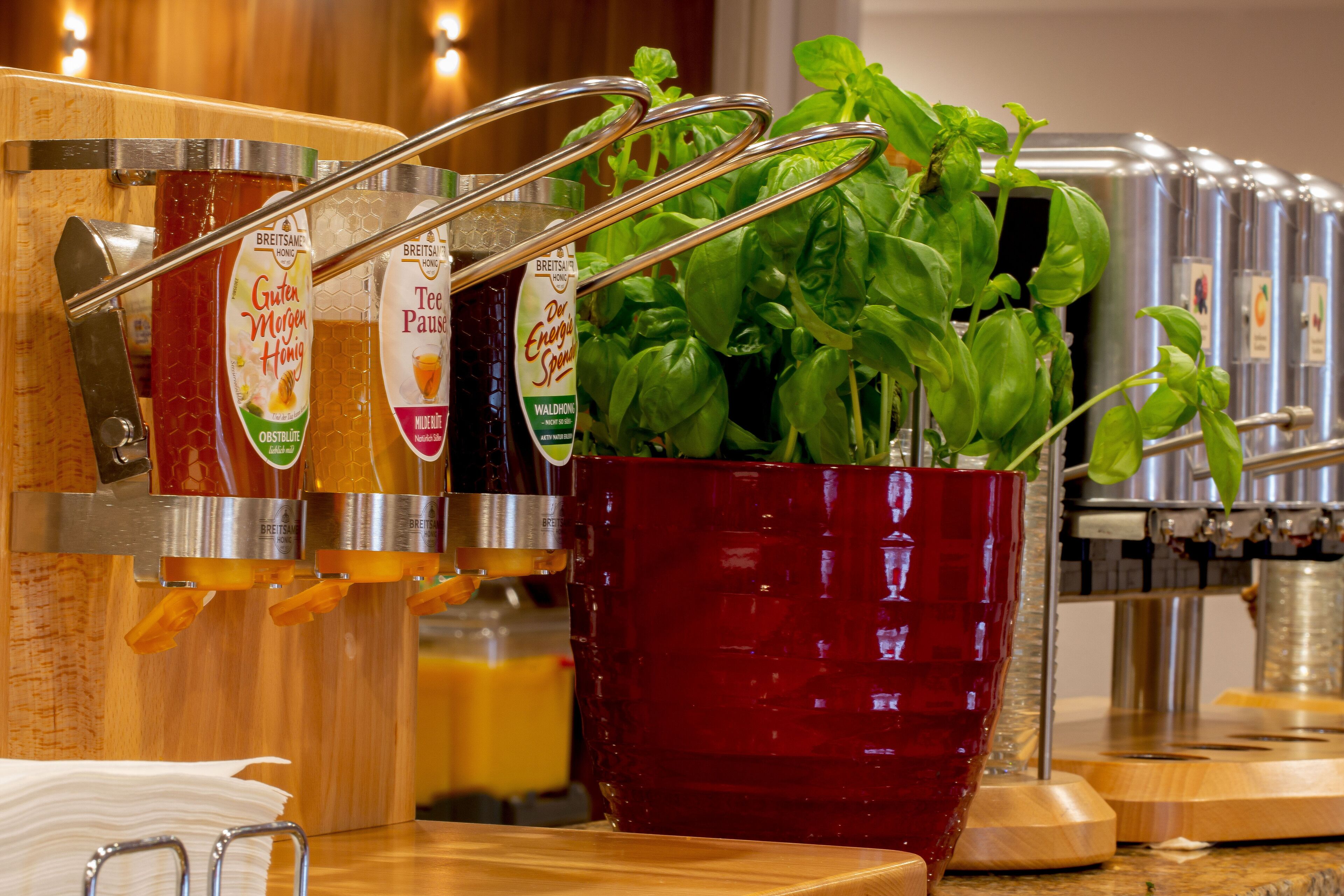 Een drankdispenser met verschillende opties, een rode pot met basilicum, en een achtergrond van roestvrij staal.